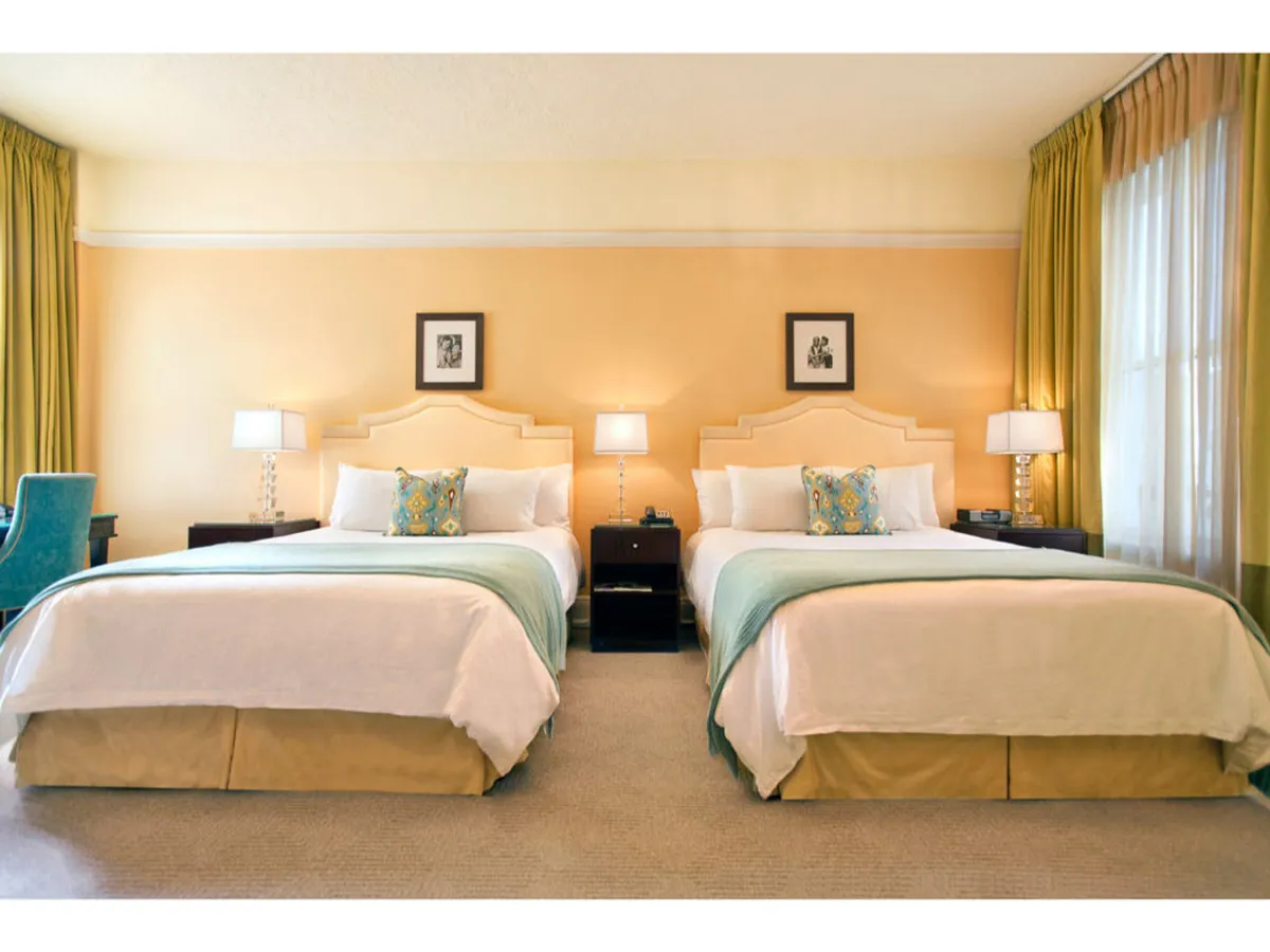 The image showcases a chic hotel suite with two plush queen beds adorned with decorative pillows, elegant nightstands with stylish lamps, soft curtains, and tasteful artwork lining the walls. The room exudes a welcoming, comfortable atmosphere perfect for relaxing after a day exploring Portland.