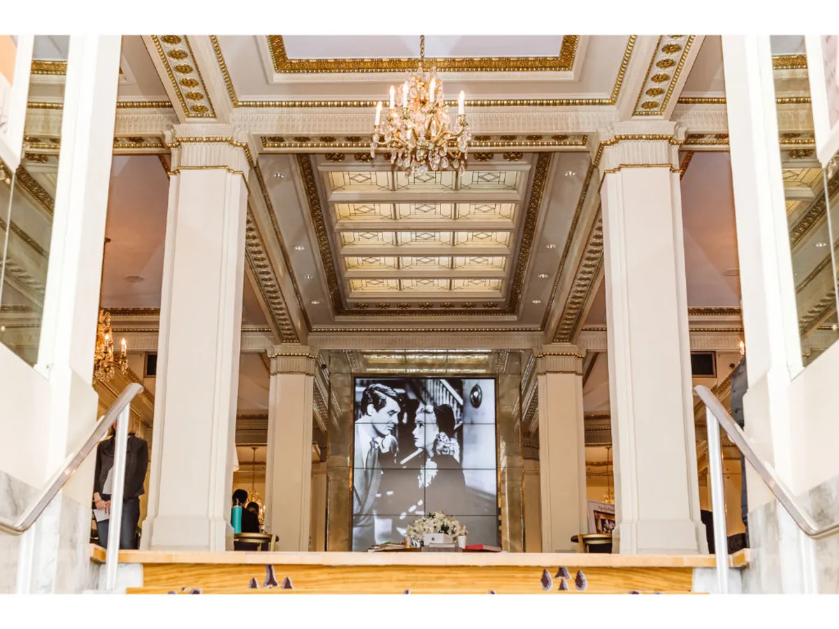 A grand, ornate interior featuring a sparkling chandelier, an elaborate ceiling, marble staircase, decorative pillars, and a large screen displaying a black and white photograph. The space evokes a sense of luxury and is set up for formal gatherings or elegant events.