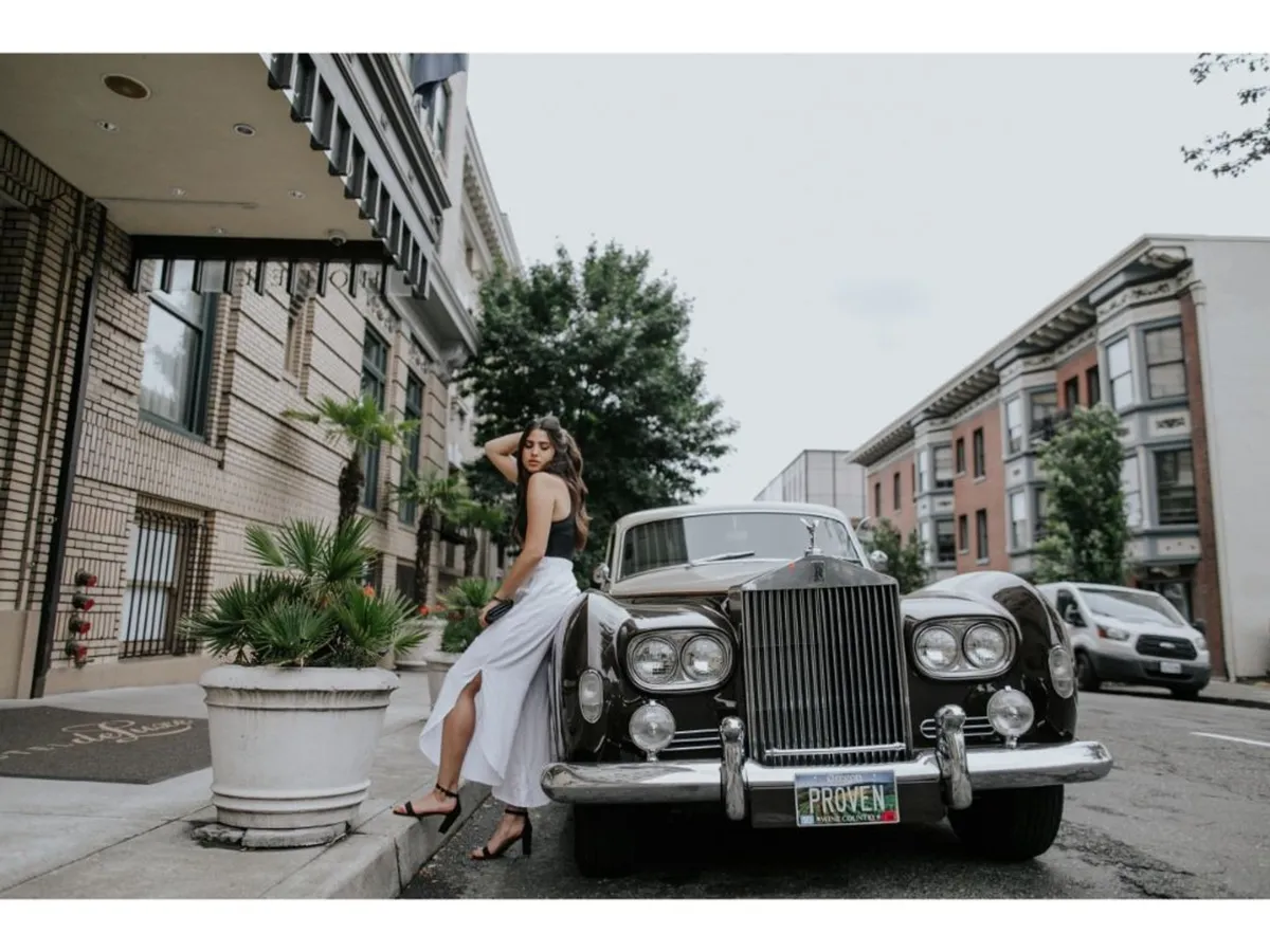 A stylish scene outside Hotel Deluxe, with a classic car parked in front and a woman in a chic white dress. The historic building’s facade sets the stage for urban exploration and contemporary style. The image conveys a sense of arrival and the blending of old Hollywood glamour with modern city culture.