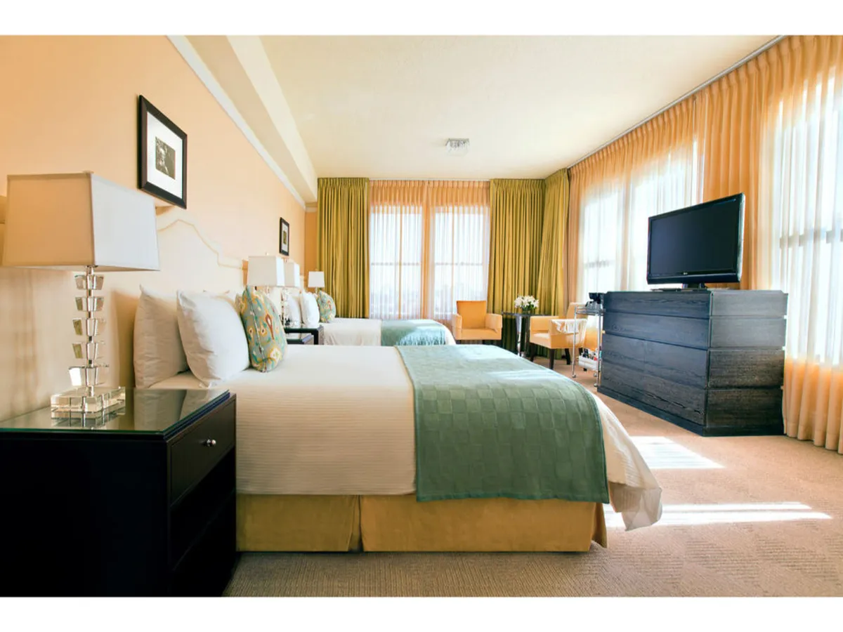 This image showcases a spacious suite featuring two neatly made queen beds situated beside large windows with sheer curtains. The room is accented by a dark wooden dresser and a comfortable seating area, with natural light filling the space and creating a tranquil atmosphere ideal for relaxation or socializing.