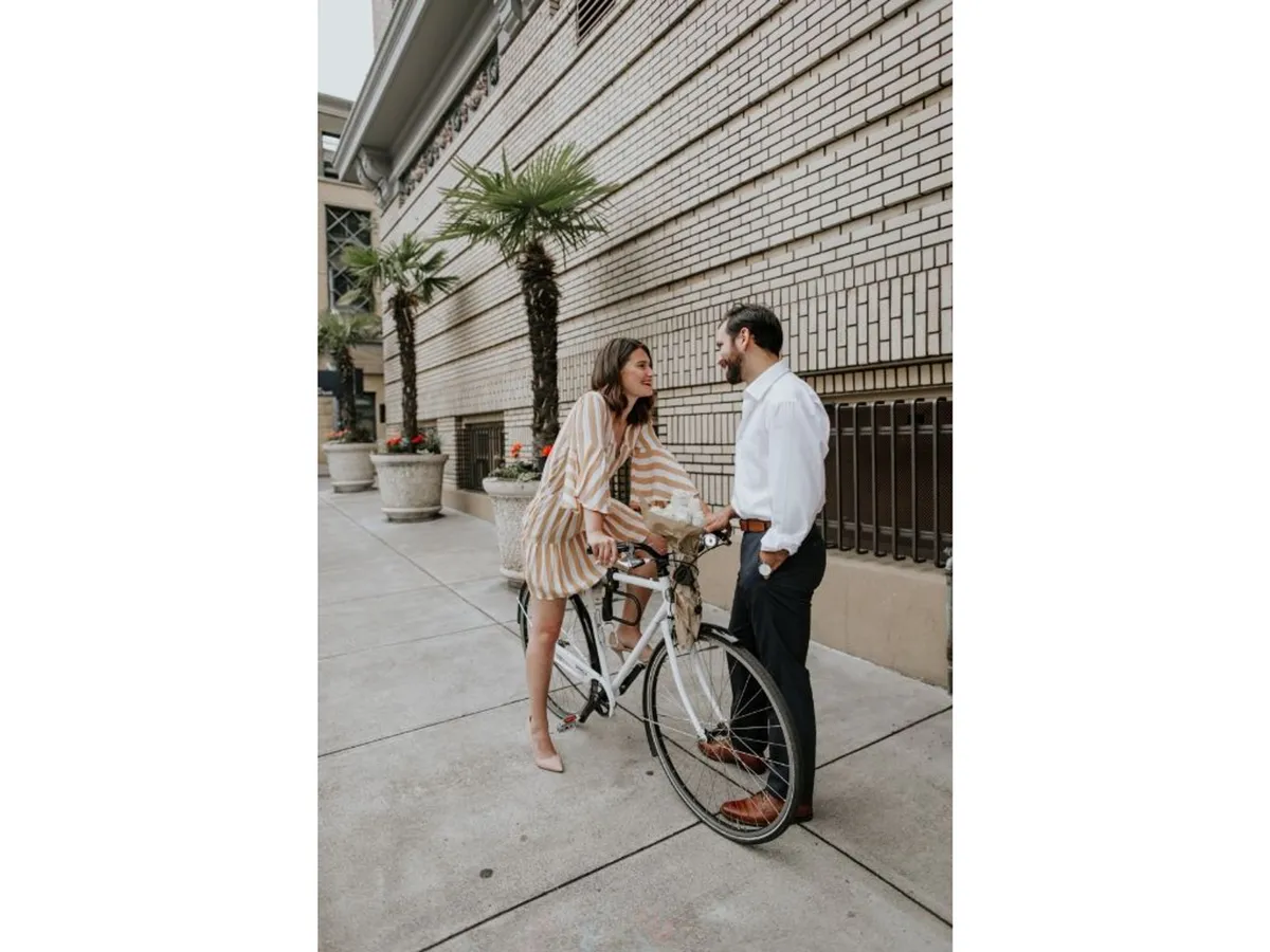 A romantic, playful moment between a couple next to a bicycle against a brick wall adorned with potted plants. The scene suggests urban exploration, casual outings, and the joy of discovering the city with a companion.