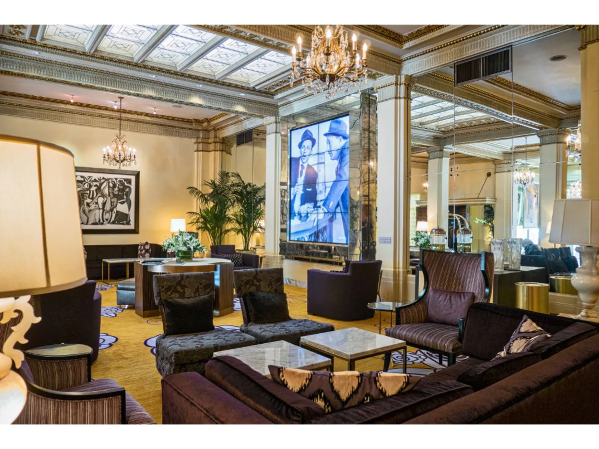 A luxurious lounge with a large chandelier, contemporary art, plush seating, and a prominent screen displaying historical imagery. The space buzzes with people socializing, networking, and engaging in group activities, evoking the feel of an upscale cinema or multi-use venue.