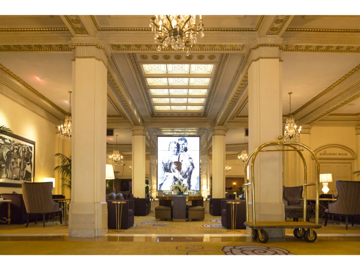 A lounge area with plush seating, dramatic chandeliers, and eye-catching large artwork sets a luxurious, inviting mood. The presence of a luggage cart hints at guest arrivals, while the seating arrangement encourages socializing and relaxation.