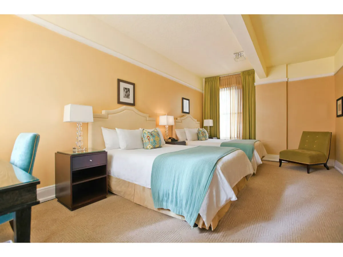 A calm and inviting bedroom scene featuring two beds dressed with turquoise throws, decorative pillows, and lamps on nightstands. A green accent chair and a writing desk offer both comfort and a dedicated workspace for productivity, while the overall setup encourages relaxation, reading, or social interaction.