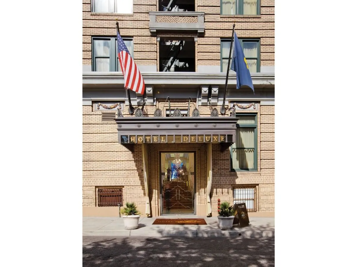 The image showcases the elegant exterior of Hotel Deluxe, featuring the grand entrance door, hotel name signage, international flags, and lush planters. The scene feels inviting and hints at the cosmopolitan, cultural atmosphere that awaits inside. The visible entrance and gathering spots suggest a seamless connection between the vibrant city outside and the luxurious experience within.
