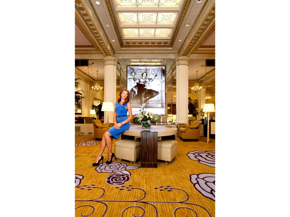 A central figure in a blue dress stands in an inviting, art-filled lounge area with a decorative ceiling and vibrant floral arrangement. The mood is lively and welcoming, perfect for socializing and relaxation after a day out.