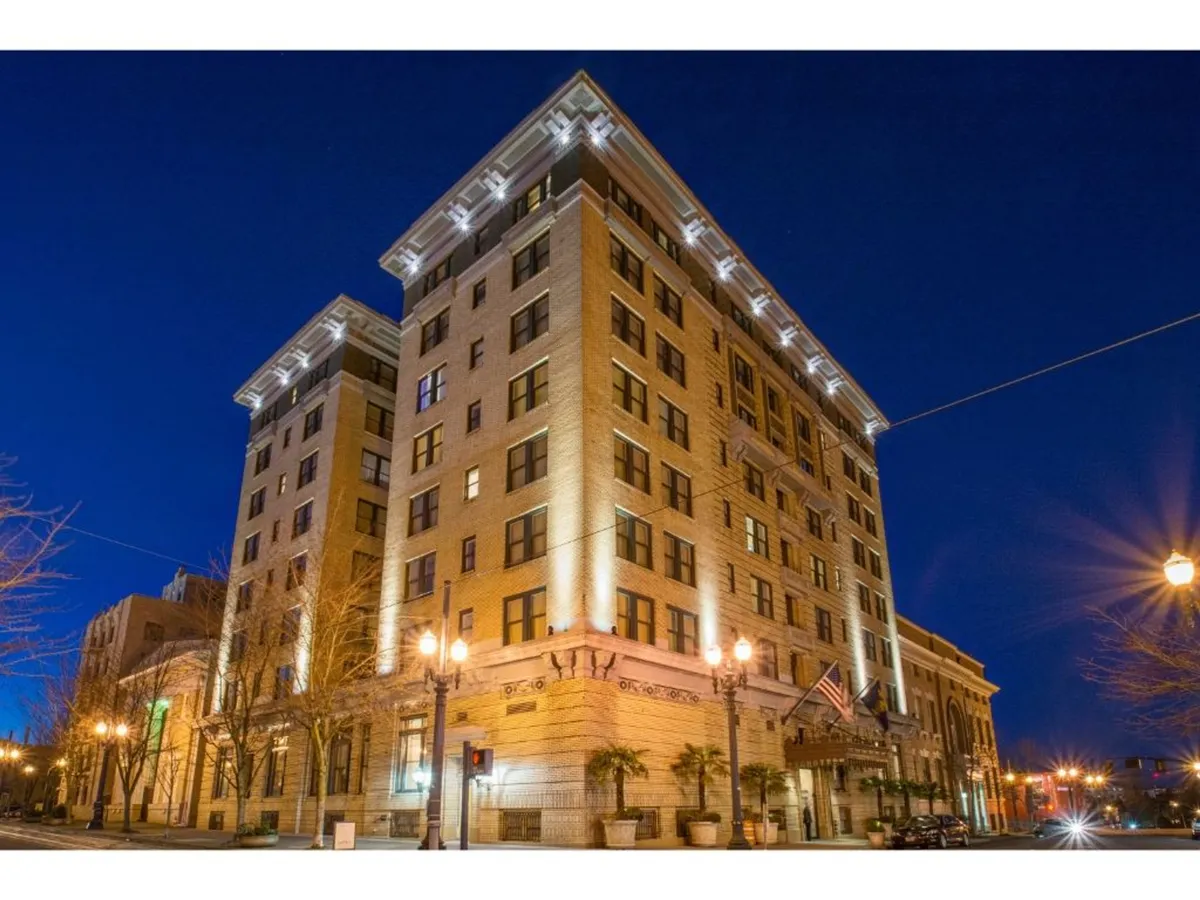 An evening exterior view of a grand building, illuminated with warm lights. Ornate architectural details, large windows, and an inviting entrance framed by decorative columns create a sophisticated city ambiance. Pedestrians can be imagined strolling by, drawn to the vibrancy of downtown nightlife.