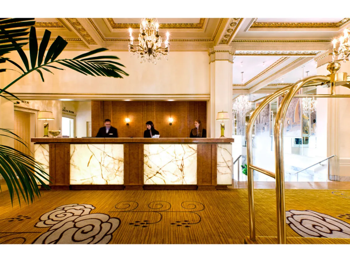 The image features the elegant reception desk with a chandelier overhead, staff present to welcome guests, and a richly patterned carpet enhancing the luxurious first impression. The setup is sophisticated with subtle touches of Old Hollywood glamour in the furnishings and decor.