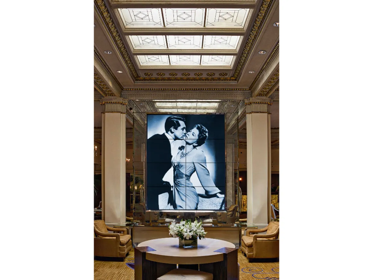 This interior scene features a luxurious lounge with a prominent black-and-white artwork of a couple kissing, a central round table adorned with fresh flowers, and plush seating arrangements. The overall ambiance is elegant and inviting, suitable for both informal meetings and relaxed socializing.